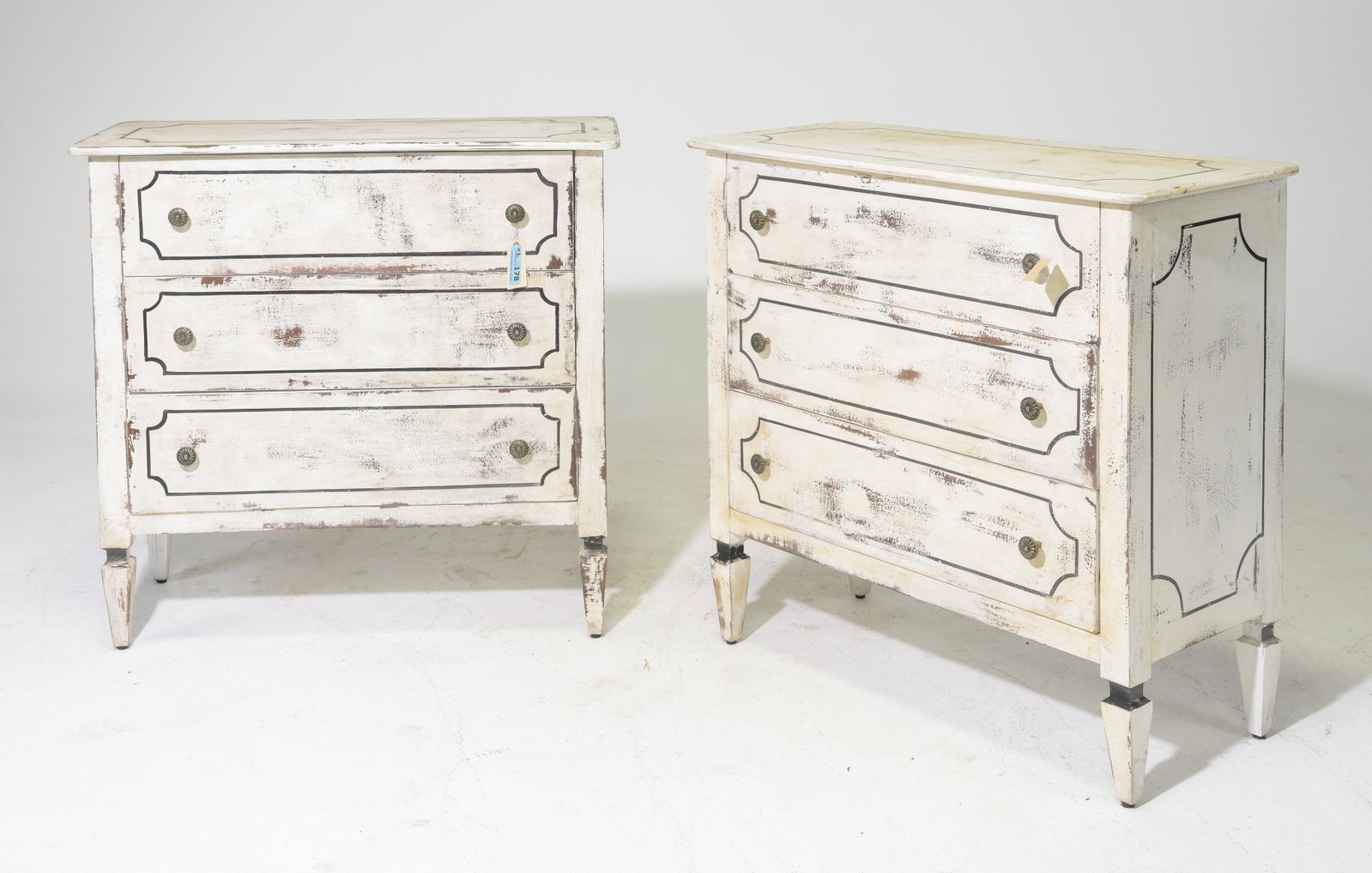 Pair Hand-Painted European Style Three Drawer Chests: A coordinating pair of French country style chests, each fitted with four drawers. The cream painted finish features black-lined raised panel decoration, dark metal rosette pulls, tapered legs, and a