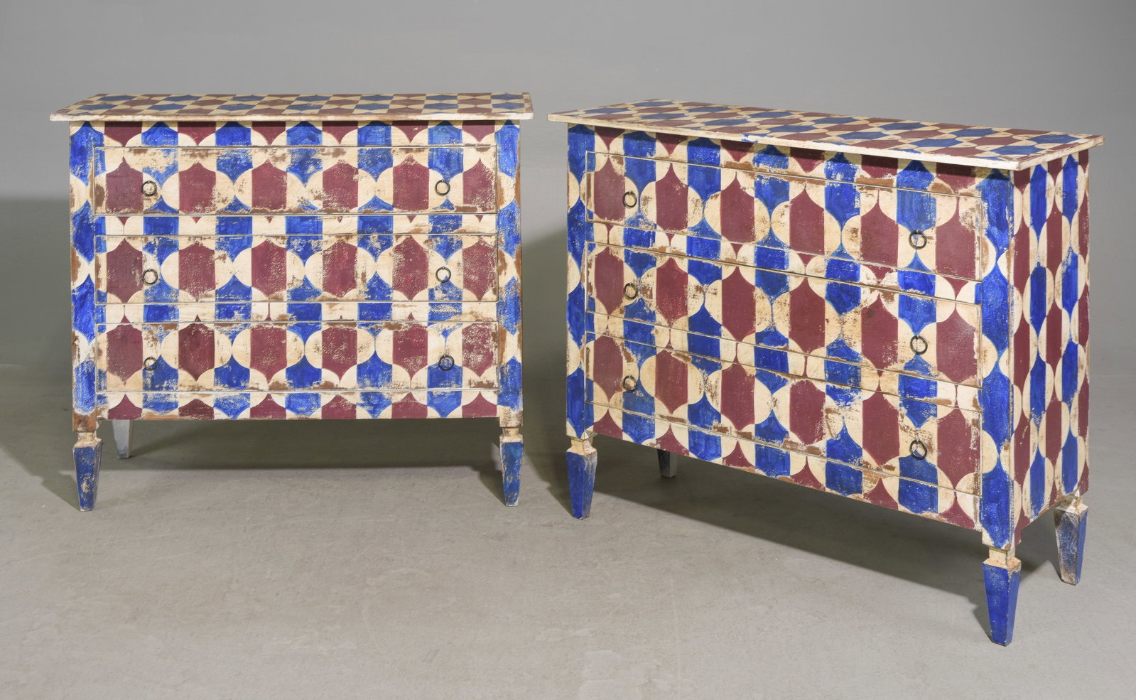 Pair Hand-Painted European Style Geometric Chests: Pair of European style three-drawer chests with bold hand-painted geometric decoration in blue, burgundy and cream. The repeating dimensional motif covers the tops, fronts and sides, with brass finish