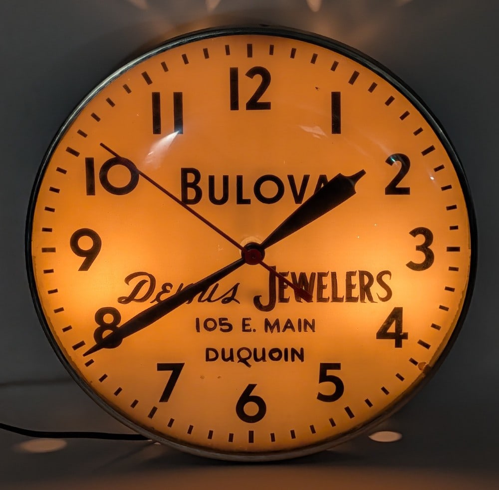 Vintage Bulova Dennis Jewelers Lighted Advertising Clock: Vintage Bulova Dennis Jewelers lighted advertising clock. The clock has some loss to the glass face and some scratches around the mounting hole on the back of the metal can. The clock lights and worke