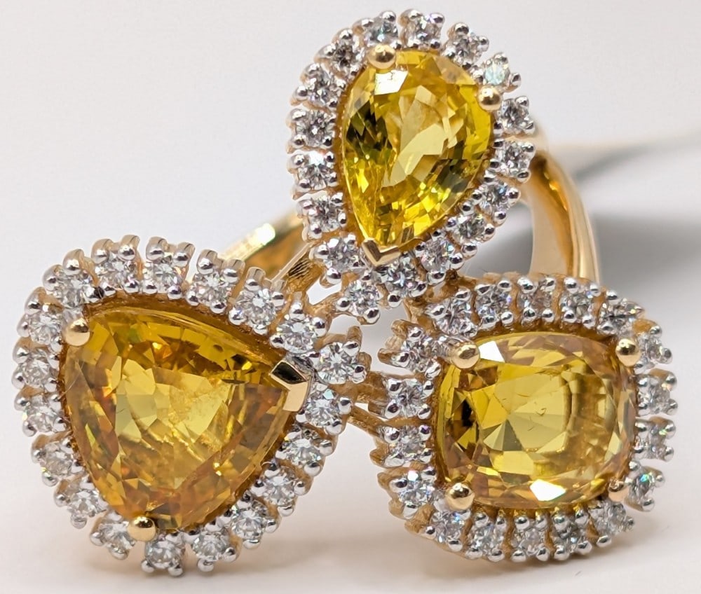 18K Gold 3 Stone Yellow Sapphire & Diamond Ring: 18K yellow gold ring with cushion, heart and pear cut yellow sapphires totaling 4.69 CT each one with a halo of diamonds totaling 0.54 CTS. Ring is size 7 and weighs 7.4 grams. Ring comes with an AIG