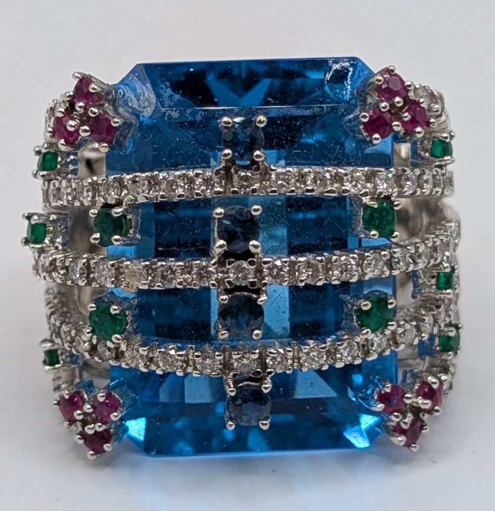 18K Gold Blue Topaz & Diamond Statement Ring: 18K white gold statement ring with large emerald cut bright blue topaz with rows of diamonds and sapphire emerald and ruby stones on top. Ring is size 6 and weighs 19.5 grams.