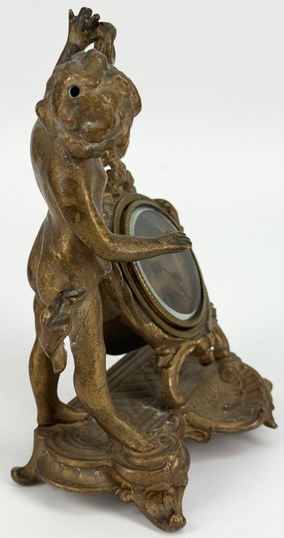 Antique New Haven Figural Cherub Desk Clock: Antique New Haven Cherub Desk Clock. The gold-finish metal clock display well and features a cherub with ribbon, face is stained. The mechanical clock is running. Measures approximately 6-1/2" tall x