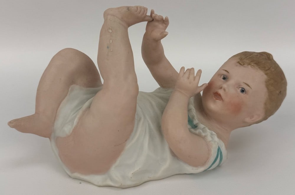 Antique Gebruder Heubach Bisque Porcelain Piano Baby: Antique Gebruder Heubach Bisque Porcelain Piano Baby. The German piano baby is laying down and holding a toe, measures 7-1/2" across. Nice condition with no chips or cracks, has some very light paint