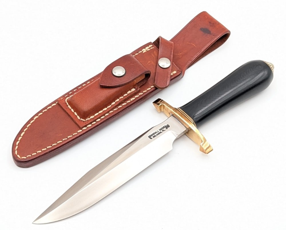 1970's Randall Model 1 7in Lugged Guard All Purpose Fighter w/ Rough Back Sheath: Features a stainless steel blade, a brass lugged double guard, a black micarta commando handle, and a scalloped brass coolie cap. Knife is in very nice condition with light scuffs and tarnishing on th