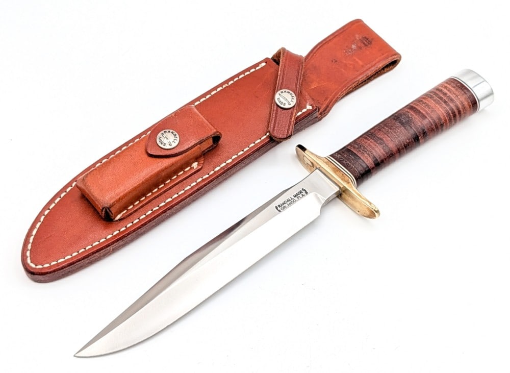 Randall Model 1 7in All Purpose Fighter w/ Sheath: Features a brass double downturned guard, a stacked leather handle, a duralumin butt cap, and a nut pommel. Knife is in very nice condition with light scuffs and tarnishing on the guard and butt cap
