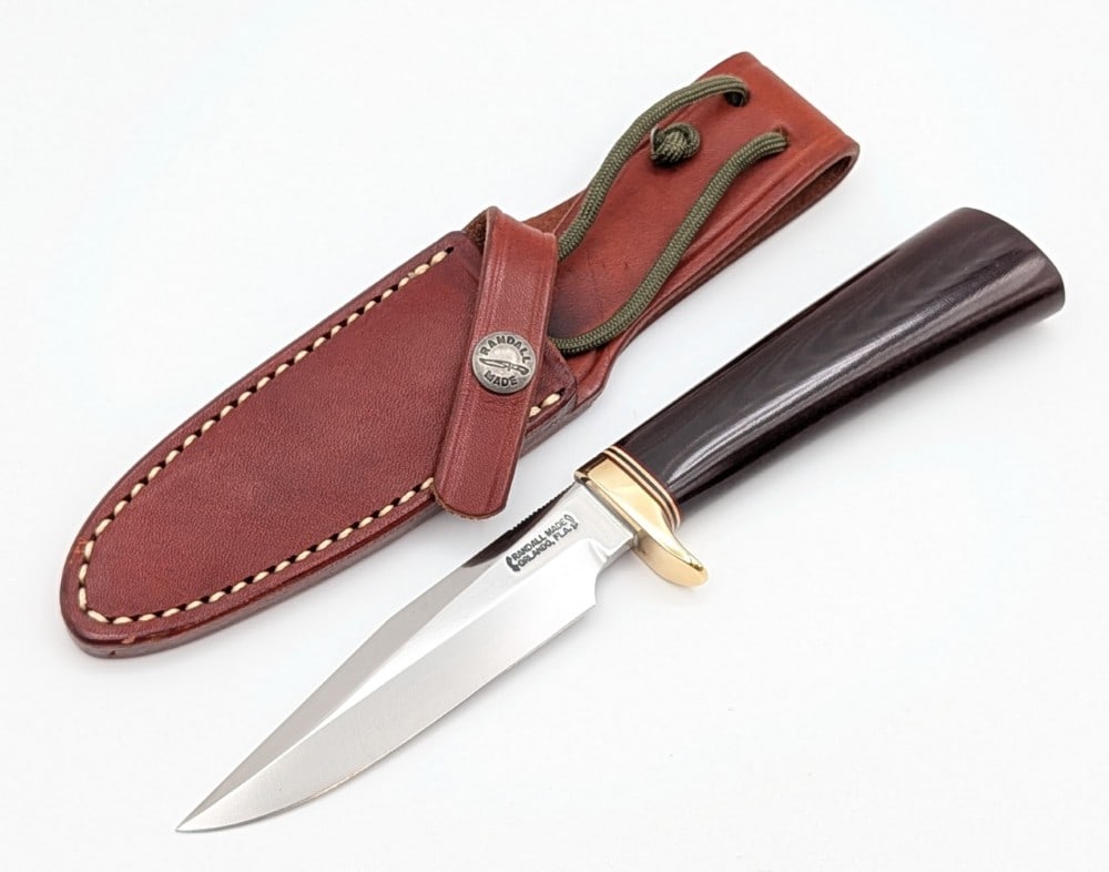 Randall Model 5 4in Camp & Trail Knife w/ Sheath: Features thumb notches on the blade, a brass single downturned guard, and a black micarta handle. Knife is in very nice condition with some possible scratch marks on the blade, and light scuffs and ta