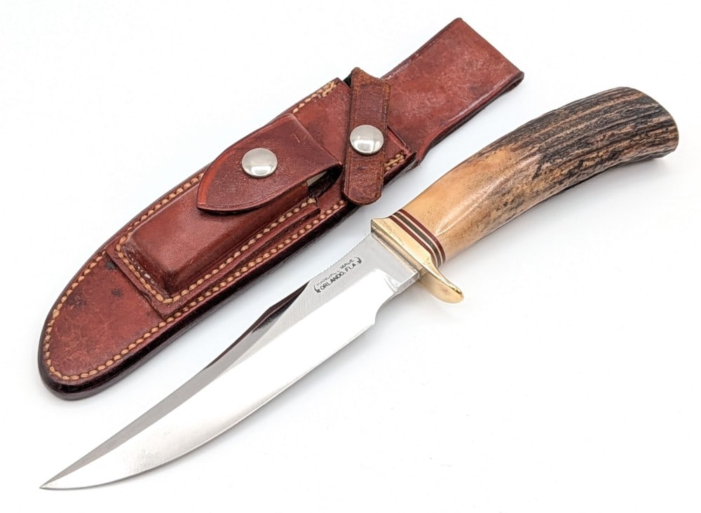 1970's Randall Model 12 6in Stag Little Bear Bowie w/ Sheath: Features a named blade with a date span Franklin Hood, thumb notches on the blade, a brass single downturned guard, and a stag handle. Knife is in good aged condition with light scratches and carbon