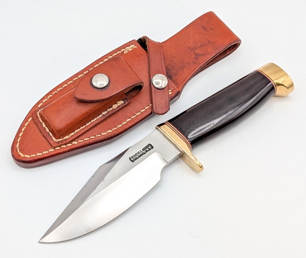 1980's Randall Model 19 5in Black Micarta Bushmaster w/ Sheath: Features thumb notches on the blade, a brass downturned single guard, a black micarta handle, and a brass crows beak pommel. Knife is in good condition with some light scratches and carbon spots on th