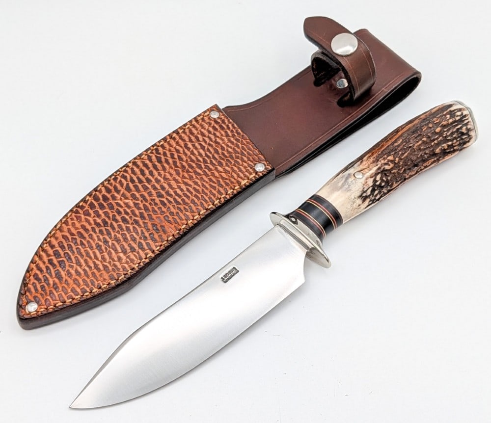 2024 Jim Behring Treeman AEB-L Pinned Stag Hunter w/ Tooled Sheath: Features a satin finish blade, a nickel silver Scagel style blade, a pinned stag handle, and a nickel silver butt cap. Knife is in like new condition. Blade length measures 5-3/4in and overall 11in.