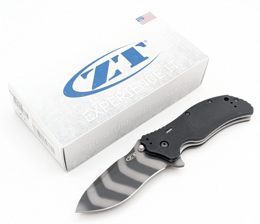 NIB Zero Tolerance ZT Model 0350 Tiger Stripe Assisted Open Folding Knife: Features a tiger stripe finish blade, thumb notches on the back of the handle, and black G-10 handle scales. Knife is in new condition. Overall measures 4-1/2in when closed.