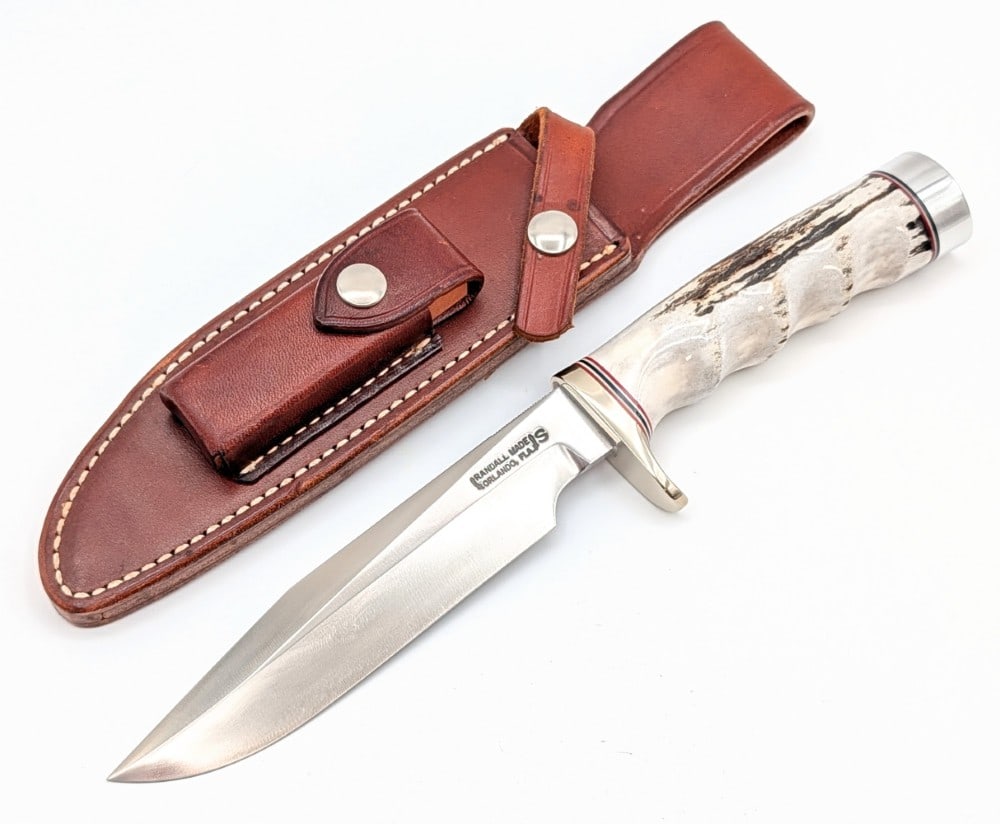1980's Randall Model 1 6in Finger Groove Stag All Purpose Fighter w/ Sheath: Features a stainless steel blade with thumb notches, a nickel silver single downturned guard, a finger groove stag handle, and a duralumin butt cap. Knife is in like new condition. Blade length measur