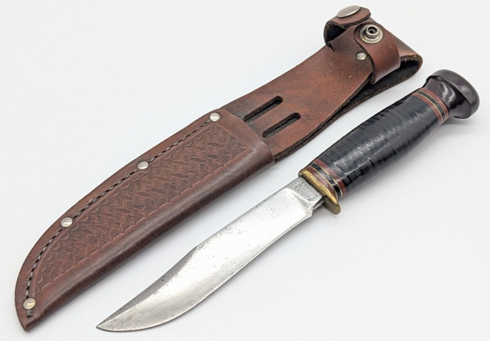 1940's-50's Marble's Ideal Hunting Knife w/ Sheath: Features a right facing serif tang stamp, a brass single guard, a stacked leather handle, and a ligume vite pommel. Knife is in used cleaned condition with material loss and scratches on the blade, an