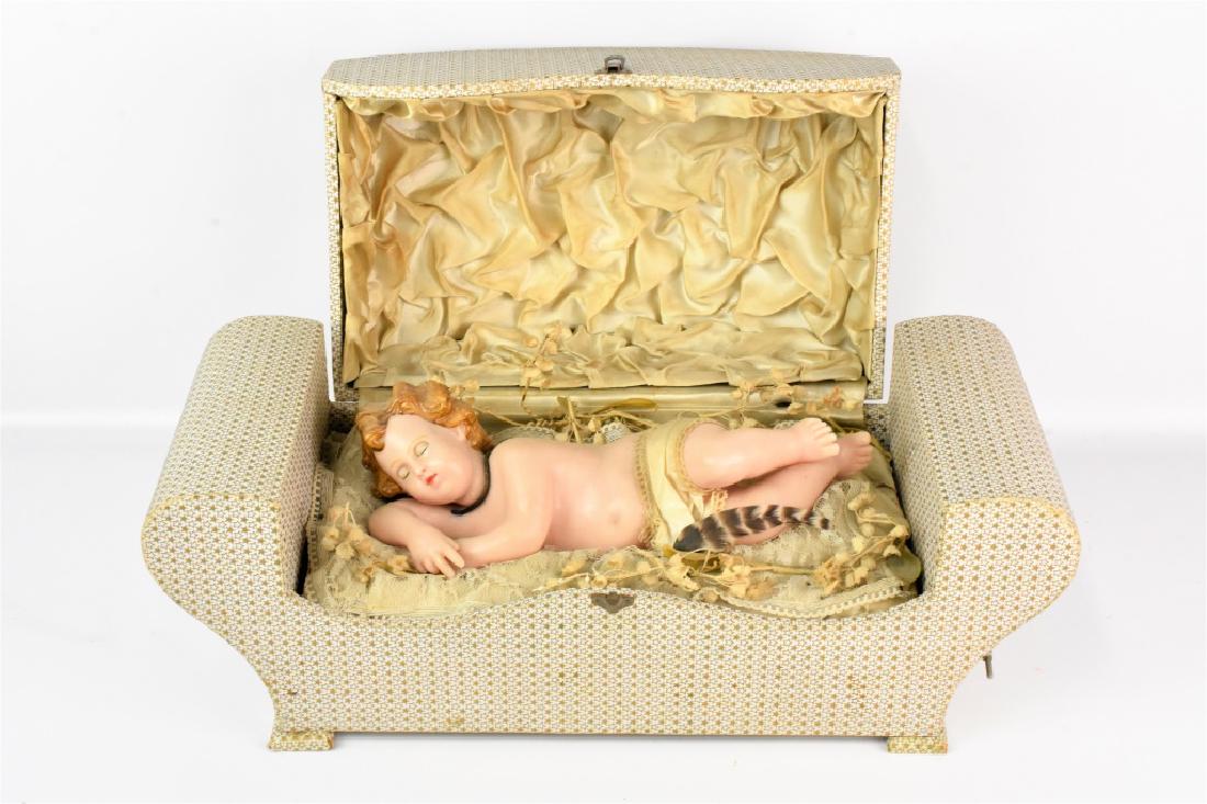 French Automaton Wax Baby Music Box: A French or Italian antique music box with automaton wax putto or baby figure possibly the baby Jesus measuring approx. 19"l by 7.5"w by 9"h. The figure rests on a lace and satin bed with faux sprigs