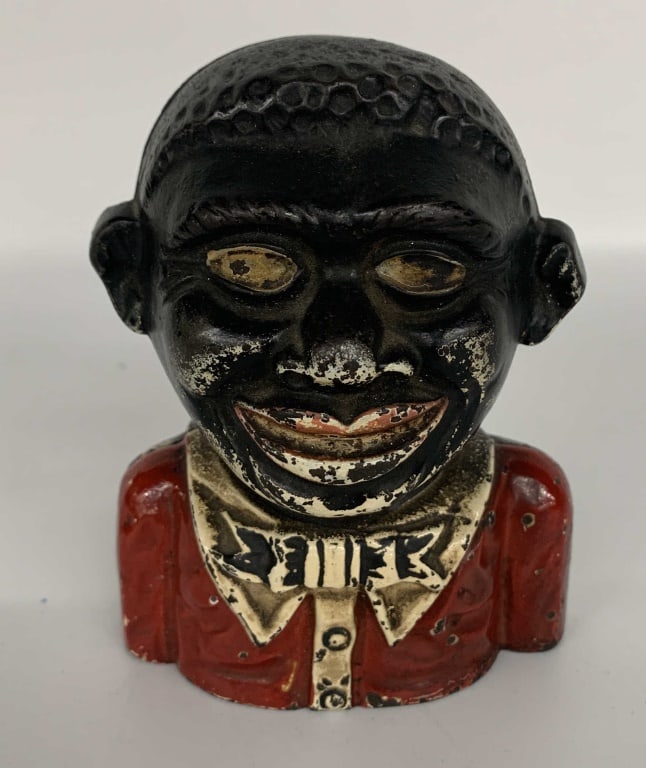 Antique Cast Iron Young "N" Still Bank: Cast iron coin bank in the form of a bust of a caricatured boy. The boy is wearing a red shirt, with white collar and black bow tie. Raised lettering on the back of the bank reads, [THE / YOUNG NIGGER
