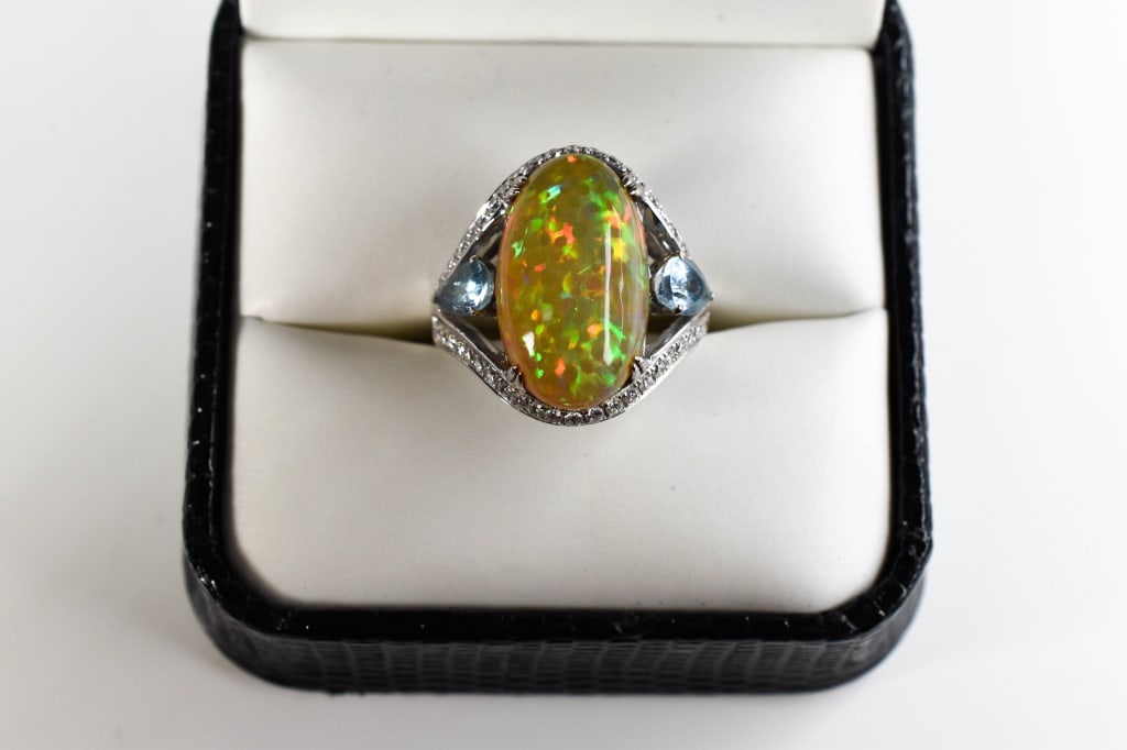 14K White Gold, Opal Cabochon & Diamond Ring: An exquisite14K white gold with large natural center opal cabochon flanked by two pear cut blue topaz and two rows of diamond accents, ring size 7, 4.87 dwt. A striking oval cabochon ring set in sleek