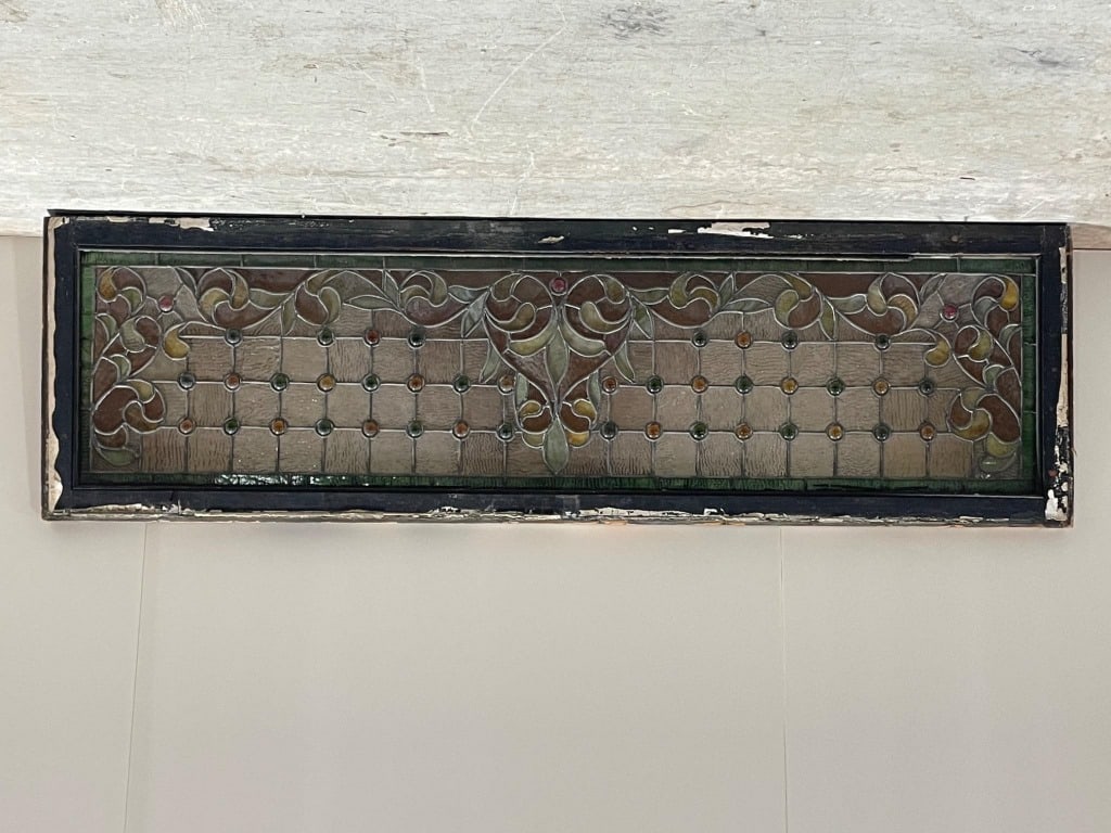 Antique Stained Glass Window with Floral Design: Stained glass window featuring a floral and geometric motif with multicolored glass accents. Secured within a wood frame, measuring 64 wide x 19.5 long. Includes a decorative arrangement of textured a
