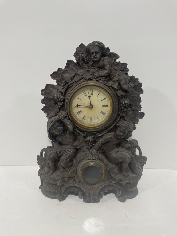 Cast Iron Mantel Clock with Figural Detail: Cast metal mantel clock featuring ornate figural details of cherubs surrounded by grapevine motifs. The clock displays Roman numerals on its face with a glass cover, accompanied by a secondary