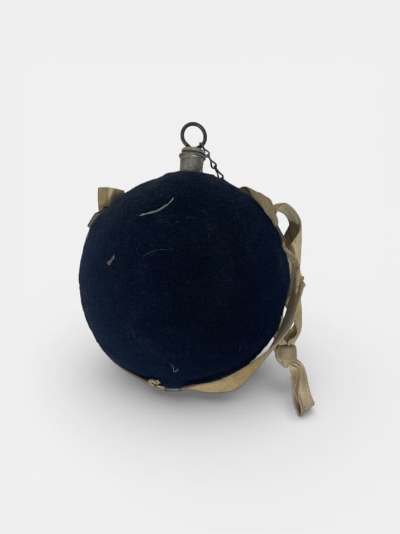 Canvas-Covered Metal Canteen: Circular canteen covered in dark canvas with a metal spout, loop, and chain. Features a fabric strap for carrying. Dimensions are 9 H x 9 W. Provenance: Acquired from a single owner private estate