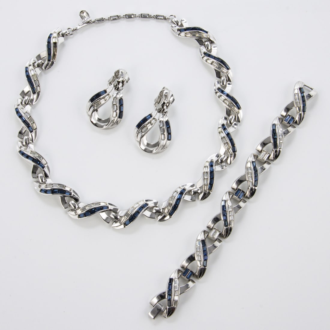 TRIFARI Alfred Philippe - Vintage Schmuckset (3 Teile): TRIFARI Alfred Philippe - Vintage Schmuckset (3 Teile)New York, um 1960-1970. Collier, Armband und Paar Ohrclips. Metall, silberfarben mit Faux-Saphiren. Collier-L.: 43 cm. B. ca. 1,5 cm. Armband-L.: