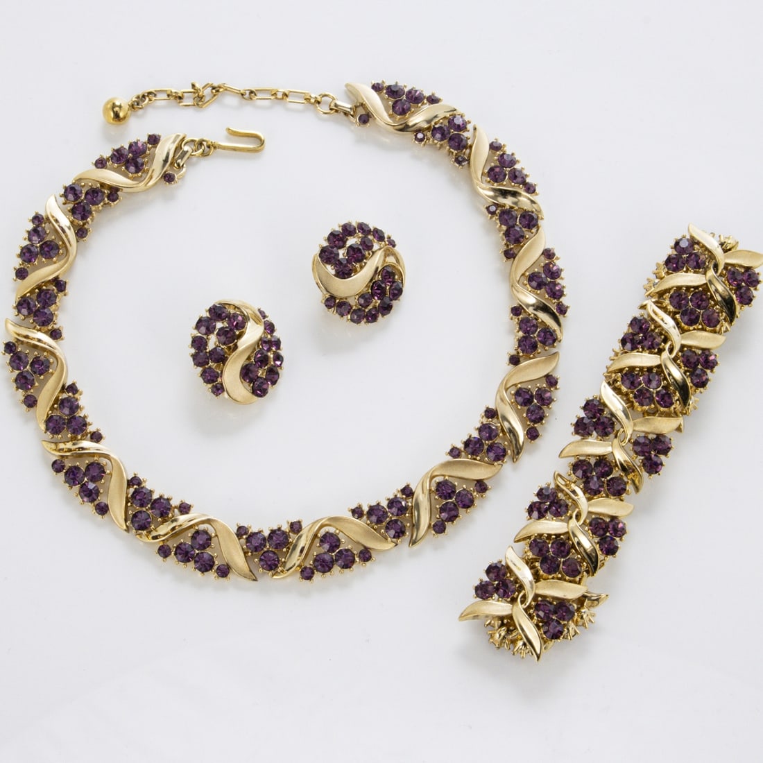 TRIFARI Alfred Philippe - Vintage Amethyst-Schmuckset (3 Teile): TRIFARI Alfred Philippe - Vintage Amethyst-Schmuckset (3 Teile)New York, um 1960 - 1970. Collier, Armband und Paar Ohrclips. Metall, goldfarben mit Faux-Amethysten-Strass. Collier-L. 31 - 37 cm. B. ca