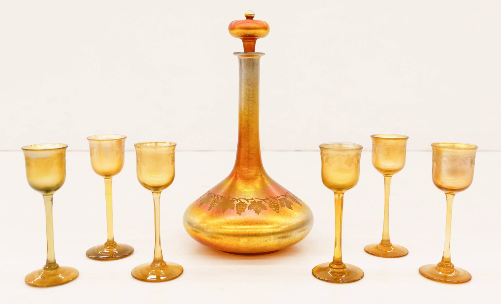 7pc L.C. Tiffany Favrile Grape Vine Decanter Set: 7pc L.C. Tiffany Favrile Grape Vine Decanter Set. Includes a gold iridescent glass stoppered bottle with intaglio cut grape vine motif 9.5''x5'' and six matching cordials 4.5''. All are signed on bott