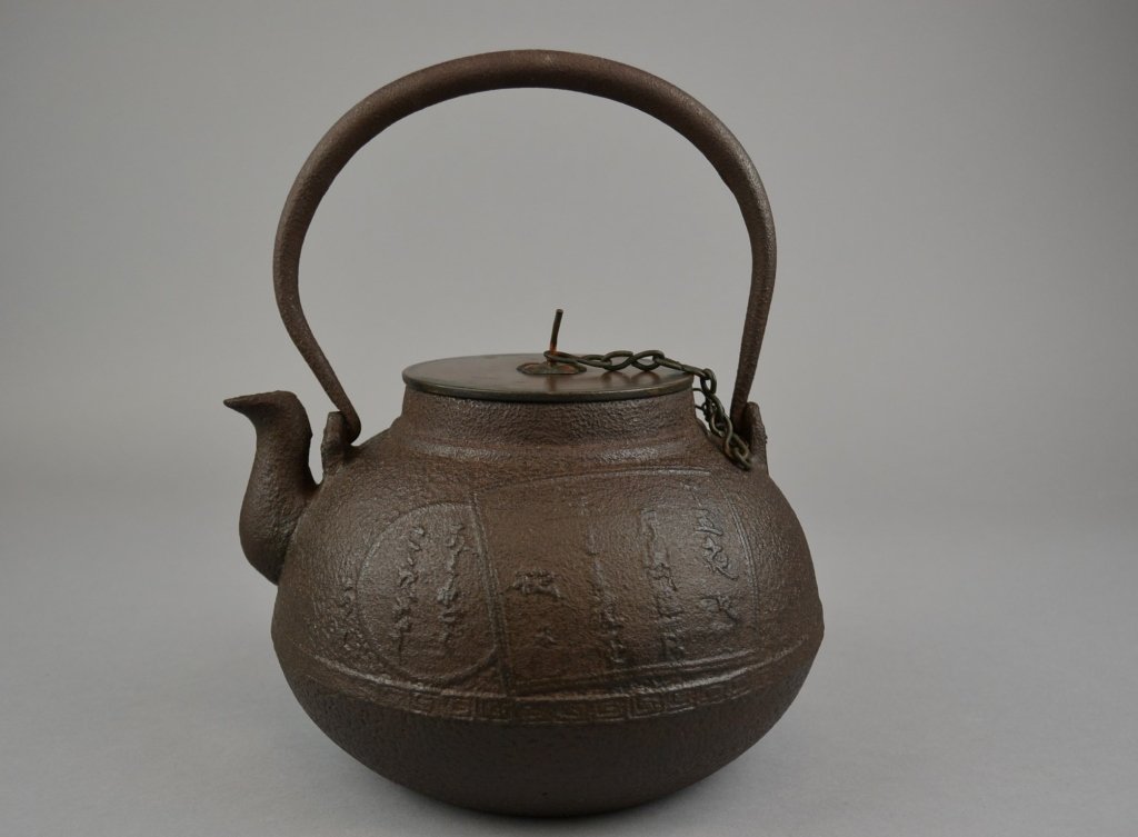 A Japanese Iron Teapot Of spherical form, one side with: Of spherical form, one side with a bird atop branches motif, the other with calligraphy medallions, with an associated bronze cover. Size: 10" height x 8 1/4" width