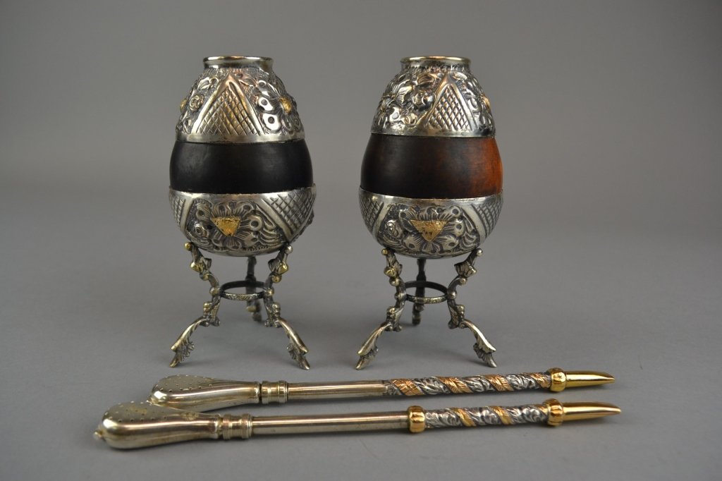 Two Argentinian Silver Bombilla and Tea Gourds Each of: Each of ovoid form, the exposed gourd flanked above and below by the chased and repousse silver mounts with gilt washed detail, marked to base and raised on three feet, and accompanied by a silver bom