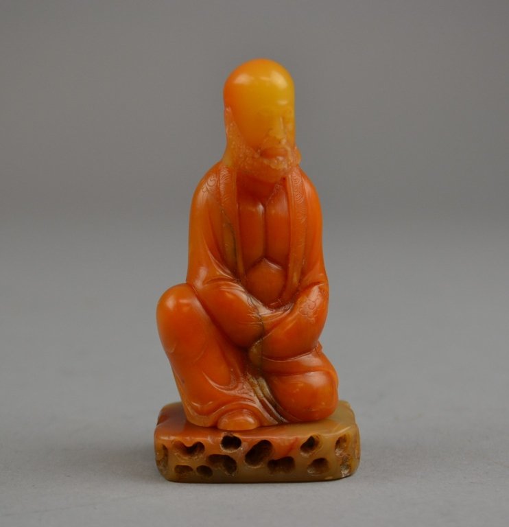 A Chinese Carved Shoushan Stone Figure Depicting a: Depicting a seated Luohan in flowing robes, having incised detail throughout, and raised on the accompanying carved soapstone base. Size: 3" height