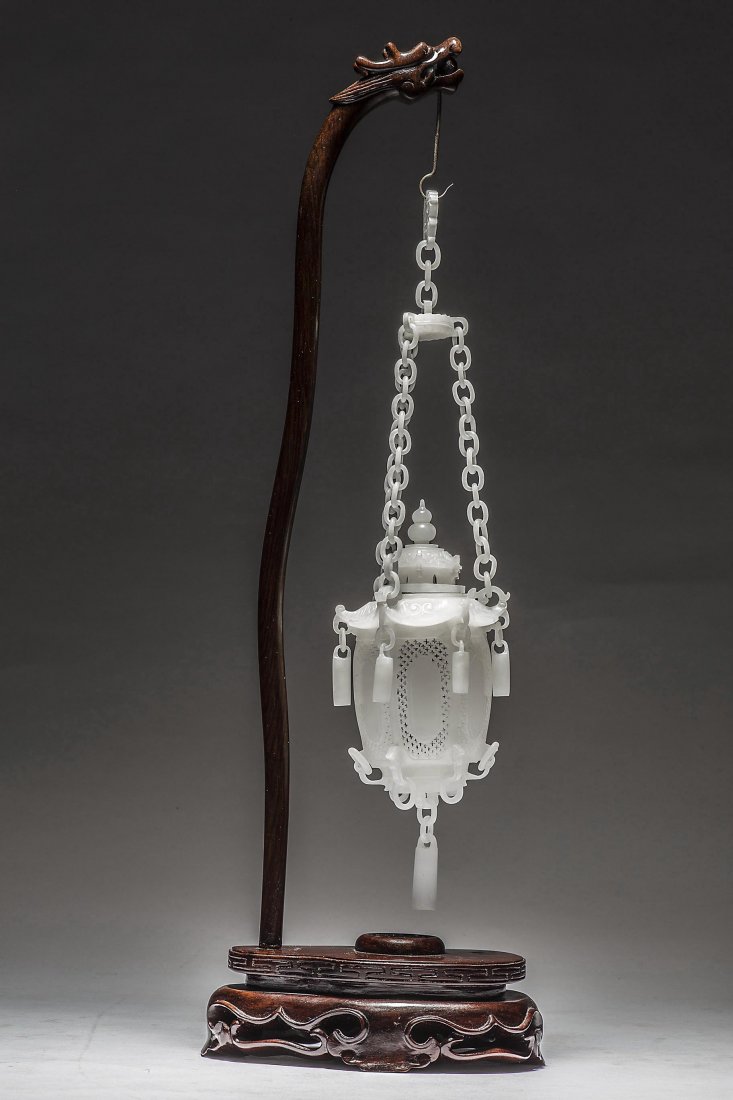 FINE WHITE JADE HANGING LANTERN: FINE WHITE JADE HANGING LANTERN The pierced body with a pagoda crest hung with tassels, suspended by a jade linked chain, on a carved dragon head wooden stand. Overall height 17 in.