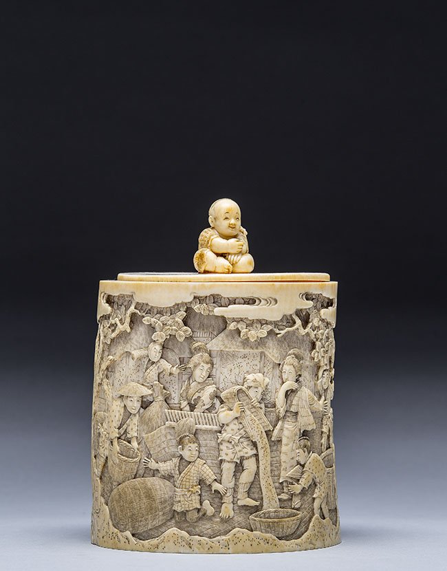 JAPANESE CARVED IVORY COVERED BOX, MEIJI PERIOD: JAPANESE CARVED IVORY COVERED BOX, MEIJI PERIOD (1868-1912) Of cylindrical form, carved in the round with figures harvesting a crop, the flat lid with a single seated boy finial, 19th century; signed.