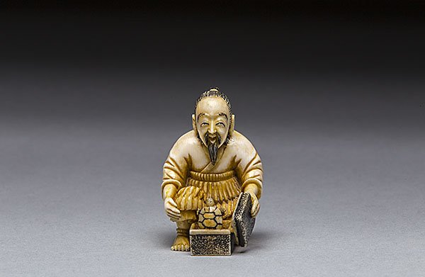 CARVED IVORY NETSUKE: CARVED IVORY NETSUKE Of a man, kneeling with a box containing a turtle and his left hand holding a book; signed. Height 2 in. (5.1 cm.)