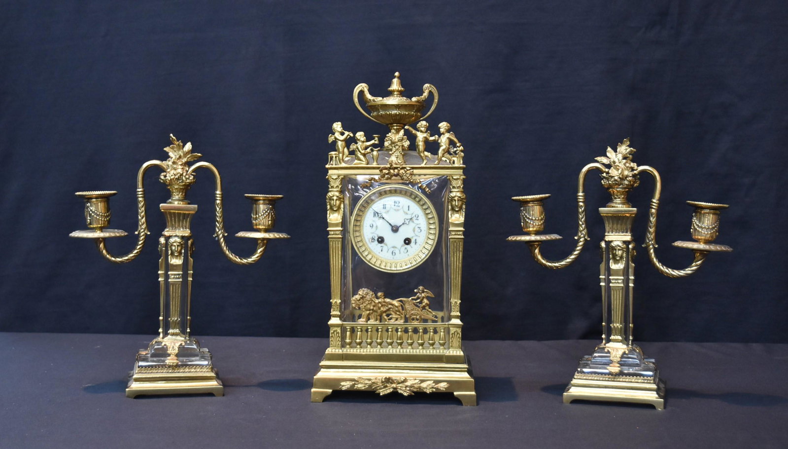 (3)pc FRENCH EMPIRE BRONZE & CRYSTAL CLOCK SET: (3)pc FRENCH EMPIRE GILT BRONZE & BACCARAT CLOCK SET WITH BRONZE CHERUB ORMOLU & BRONZE CHARIOT , TRIPLE CANDLESTICKS HAVE FEMALE MASKS - CLOCK IS 7" x 6" x 15" ; CANDLESTICKS ARE 11 1/2" TALL