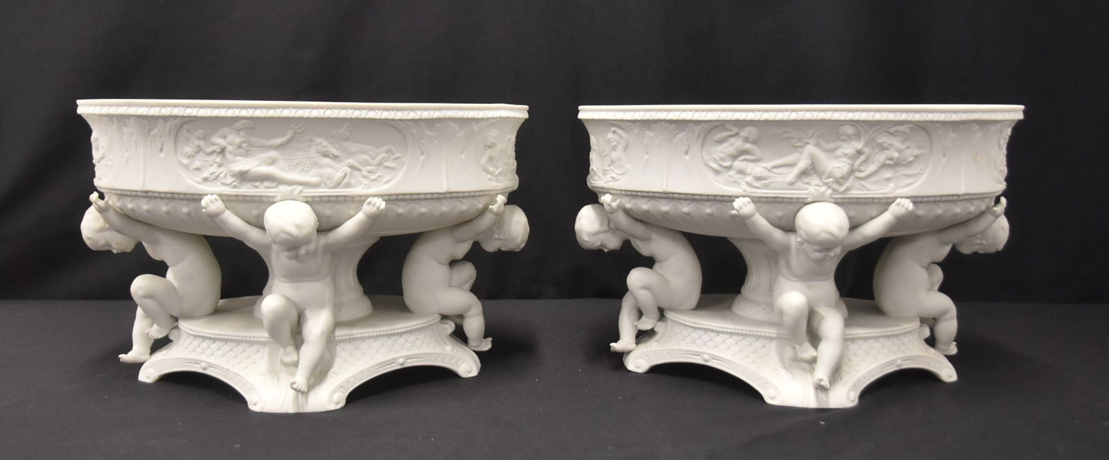 PAIR PARIAN BISQUE PORCELAIN CENTERPIECES: (Pr) ANTIQUE PARIAN BISQUE PORCELAIN CENTERPIECE PEDESTAL BOWLS WITH PUTTIS HOLDING BOWL SURROUNDED BY MEDALLIONS OF MYTHOLOGICAL SCENES - 14" x 10" x 9"
