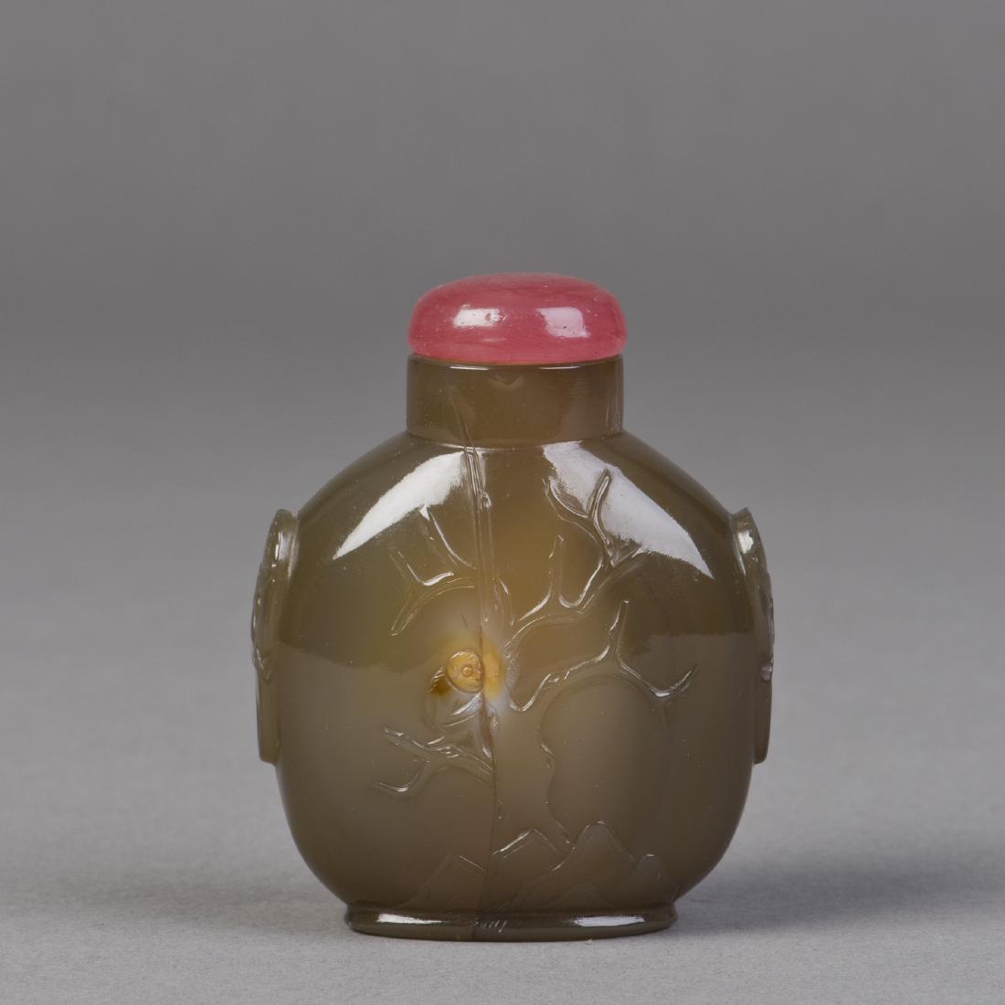 AN AGATE SNUFF BOTTLE: A snuff bottle made of agate featuring light relief of leafless tree and rocks on the front and relief handles of lion form, includes a red cap. Height 3" Width 2"