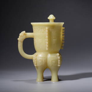 A CHINESE YELLOW JADE TAOTIE MASK WINE VESSEL JUE: The vessel is cast standing on two tapering legs. Its body is divided into three deep lobes , everted rim and one handle. The body is flat-cast decorated with three large round-horned taotie masks