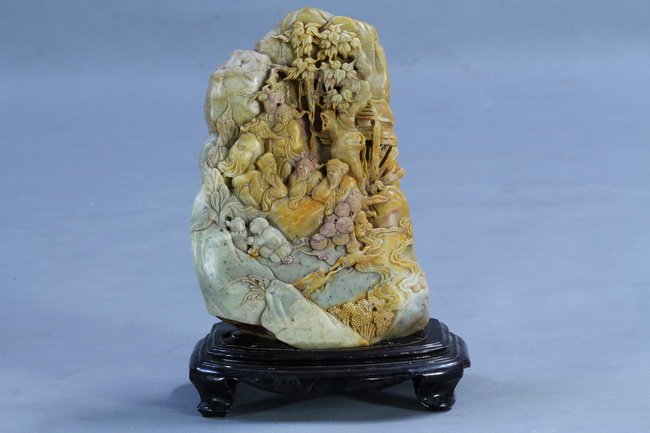 Chinese Carved Soapstone Shoushan: Carved soapstone mountain with figural and landscape scene with wooden stand. Irregular stone of beige tone with caramel inclusions, China; unmarked. Length 7" Height 13"
