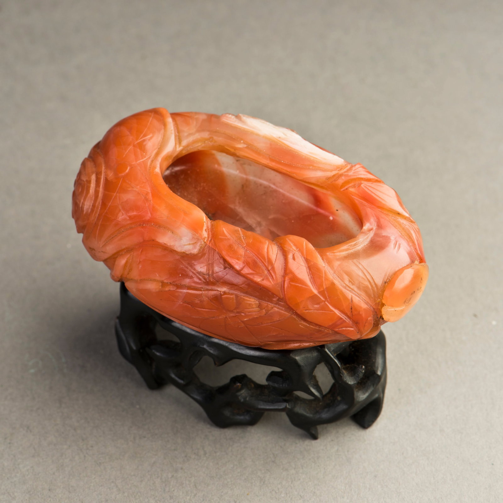 NANHONG AGATE BRUSH WASHER WITH STAND: Nanhong agate brush washer with stand. The brush washer is carved from an oval piece of Nanhong agate with a foliage motif in shallow relief and displayed on an openwork wooden stand. 0.98"h x 2.17"w