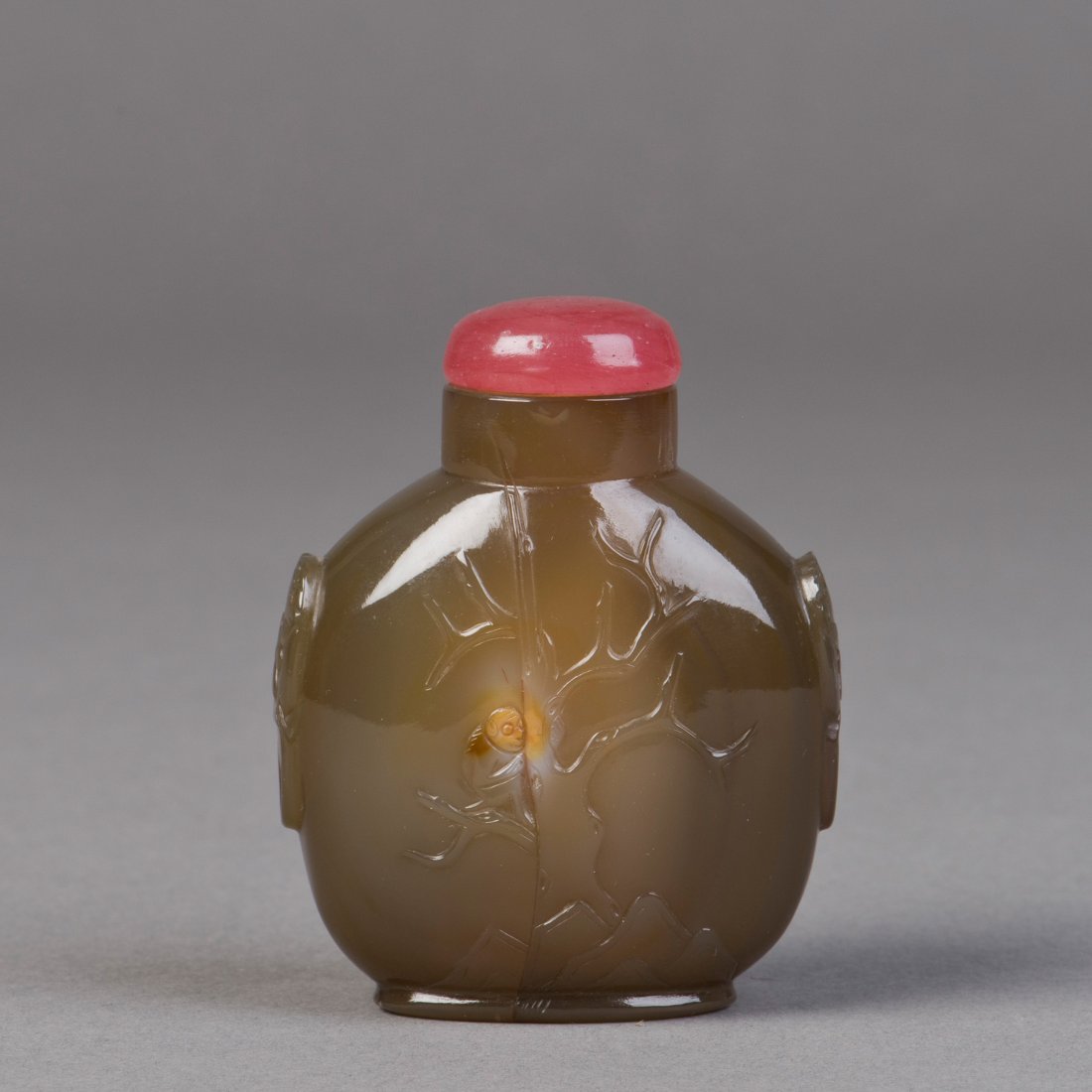 AN AGATE SNUFF BOTTLE: A snuff bottle made of agate featuring light relief of leafless tree and rocks on the front and relief handles of lion form, includes a red cap. Height 3" Width 2"