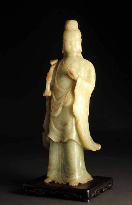 AN ANTIQUE LARGE CARVING OF A STANDING GUAN YIN CHIN: AN ANTIQUE LARGE CARVING OF A STANDING GUAN YIN CHINA Featured standing, grasping a ruyi scepter in her right hand; jade, jadeite or hardstone. Wooden stand included. Provenance : From an old Montreal