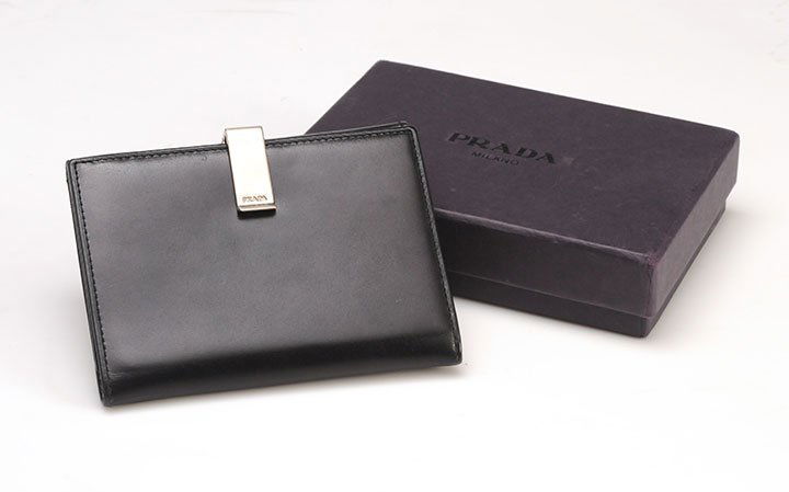 PRADA: Black leather wallet, 5 credit card slots, 1 large banknote slot and a coin compartment. Silver tone metal plier type fastener. With box.