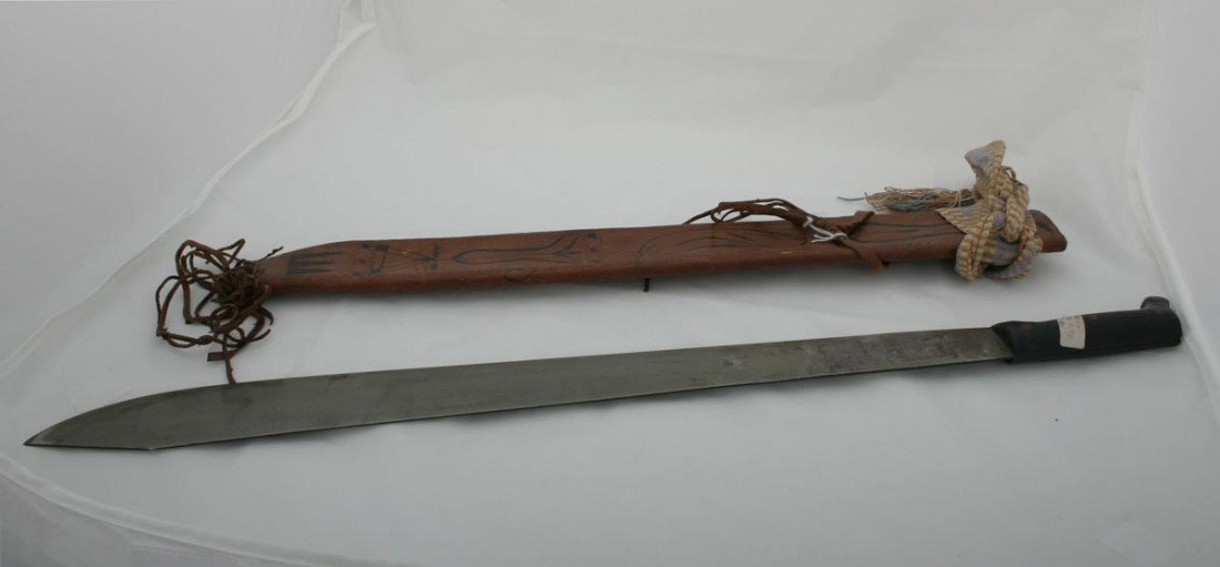 Machete: Breite Klinge, Holzgriff, Scheide aus braunem Leder bemalt. Leder-Schmuckbehang. A-Technik: Eisen. Scheide aus Leder. A-Groesze: L.: 71 cm. A-Zeit-Leben: Indonesien.