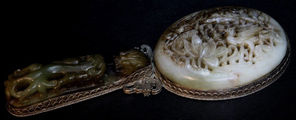 A Silver Mirror Mounted with Celedon Jade: The silver mirror mounted with two pieces of celedon jade objectives as decorations, one jade belt-hook carved with dragon and the other round jade carved with birds in a lotus pond. Length:9.5, Width