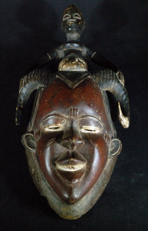 An African Carved Wood Mask: Athe tribe, depicting a stylized human face with horns, surmounted by a fetish figure. Size: 17" L