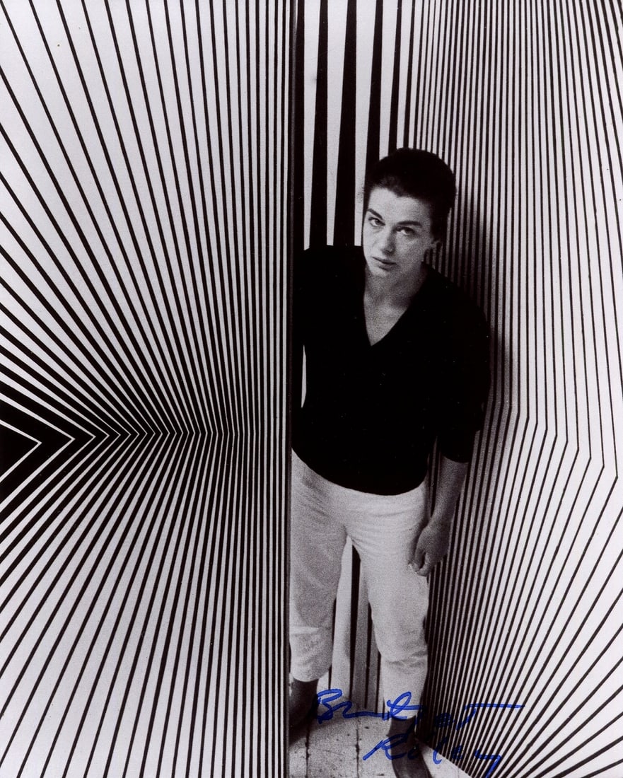 RILEY BRIDGET: (1931- ): RILEY BRIDGET: (1931- ) English Painter, known for her Op Art. A fine signed 8 x 10 photograph by Riley, the artistic image showing the artist standing in a full- length pose surrounded by several of