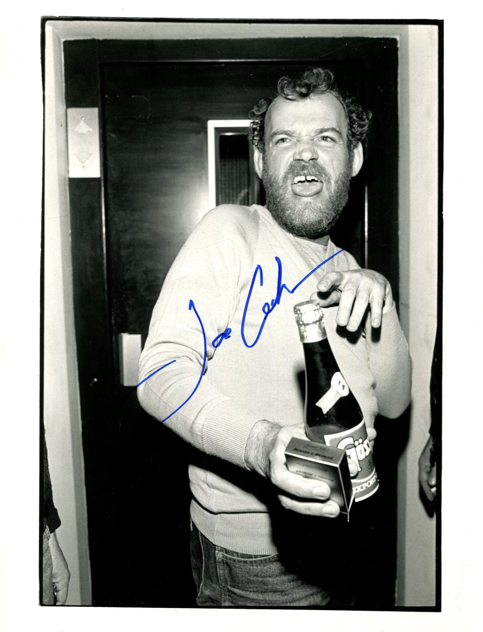 COCKER JOE: (1944-2014): COCKER JOE: (1944-2014) English Singer. Signed 6,5 x 8,5 photograph of Cocker standing in a half-length pose with an intense facial grimace, holding a bottle of beer in one hand alongside a pack of