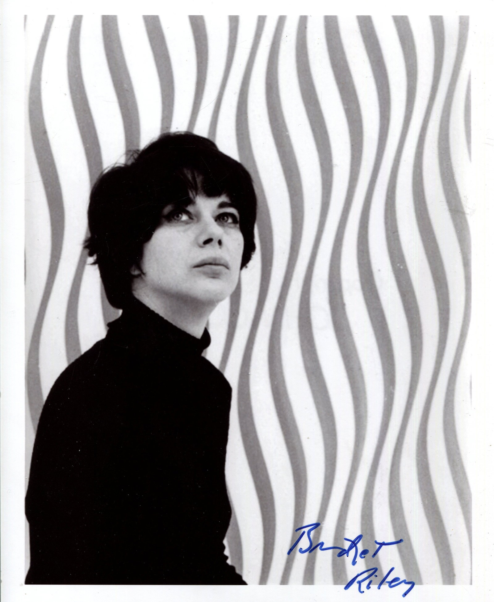 RILEY BRIDGET: (1931- ): RILEY BRIDGET: (1931- ) English Painter, known for her Op Art. A fine signed 8 x 10 photograph by Riley, the image showing the artist in a half-length pose before one of her works. Signed in bold