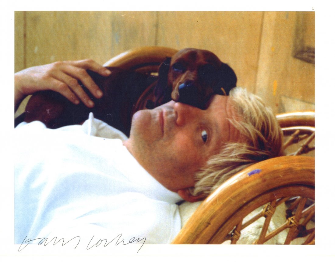 HOCKNEY DAVID: (1937- ) British Artist. Signed: HOCKNEY DAVID: (1937- ) British Artist. Signed colour 11 x 8.5 printed paper photograph, the image depicting Hockney lying in a head and shoulders pose with a small dog affectionately lying on his fac