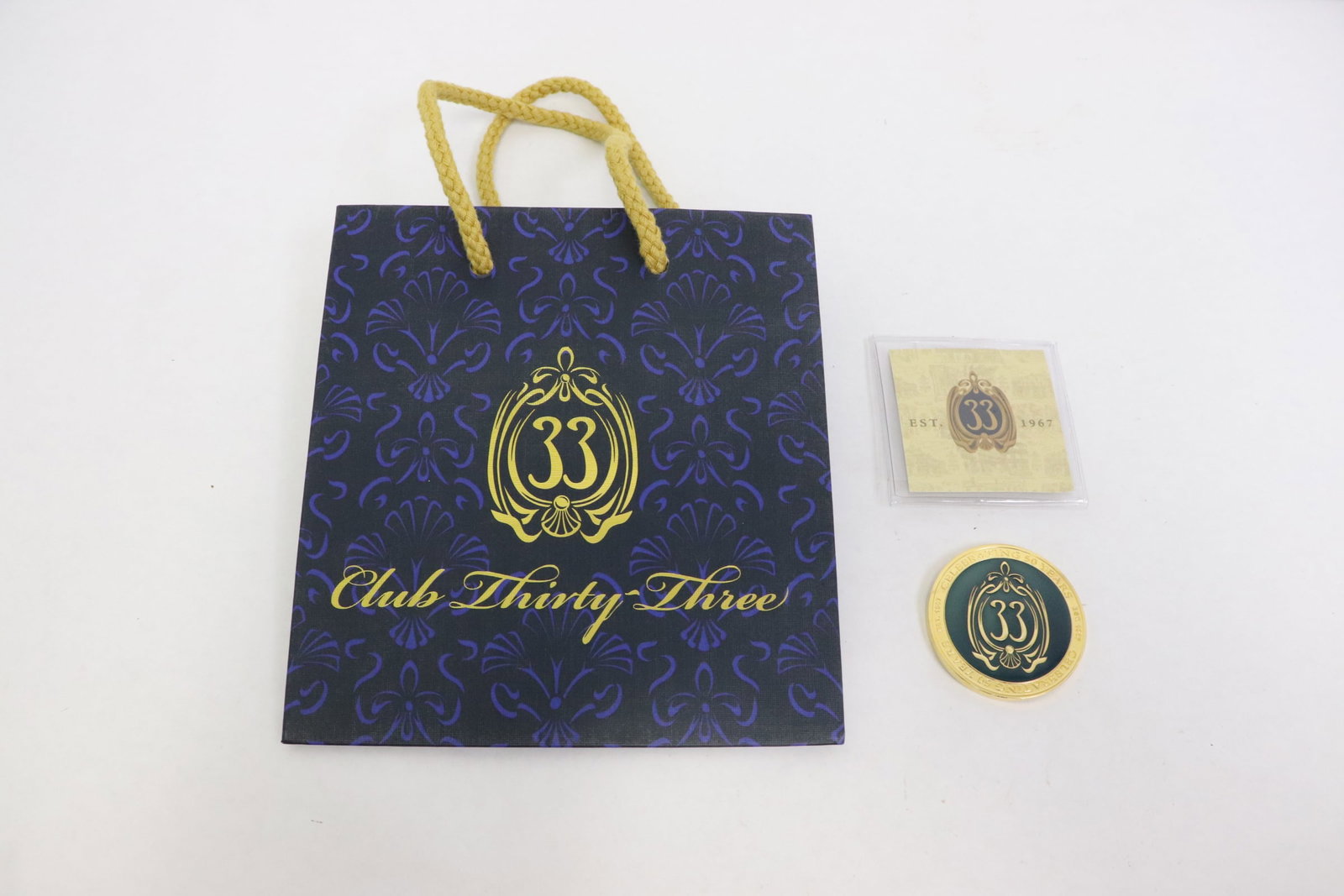 club 33 challenge coin in original sleeve, w/ a gift bag: A club 33 enamel gold tone challenge coin (0.15"x1.84"dia) issued in 2017 for 50th anniversary, one side features club 33 logo with blue enamel, the other side a gold tone club 33 logo, the coin is in