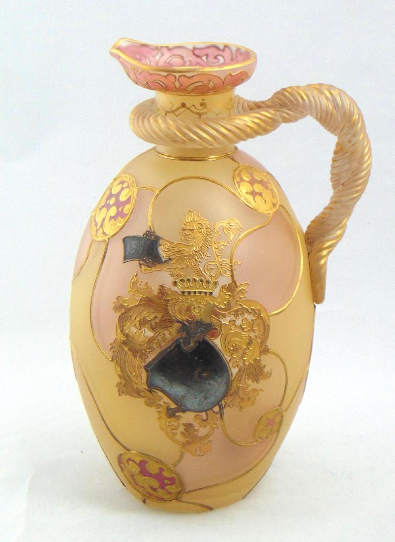 Mt. Washington Rope Handled Ewer: Cylindrical form with scrolling earth tone panels decorated with a central heraldic crest of a rampant lion, highlighted with pin wheel medallions and adorned with a rope twist handle, 10" ht. Conditi