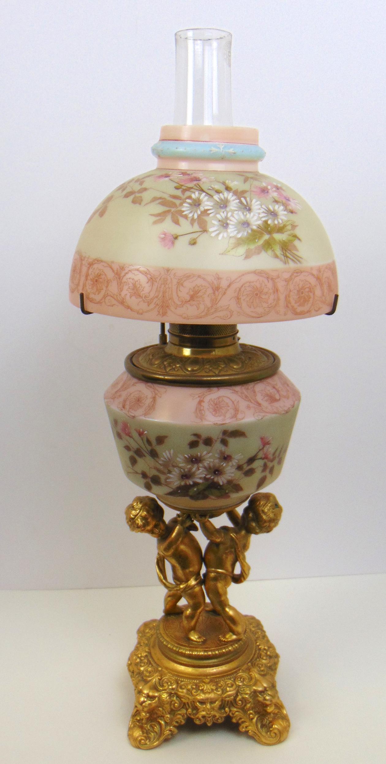 Victorian lamp with cherub base: Mt. Washington parlor lamp with a daisy decorated shade and font set on a double cherub gilt base, 27" to the top of the chimney. Condition: roughness around the top of the shade.